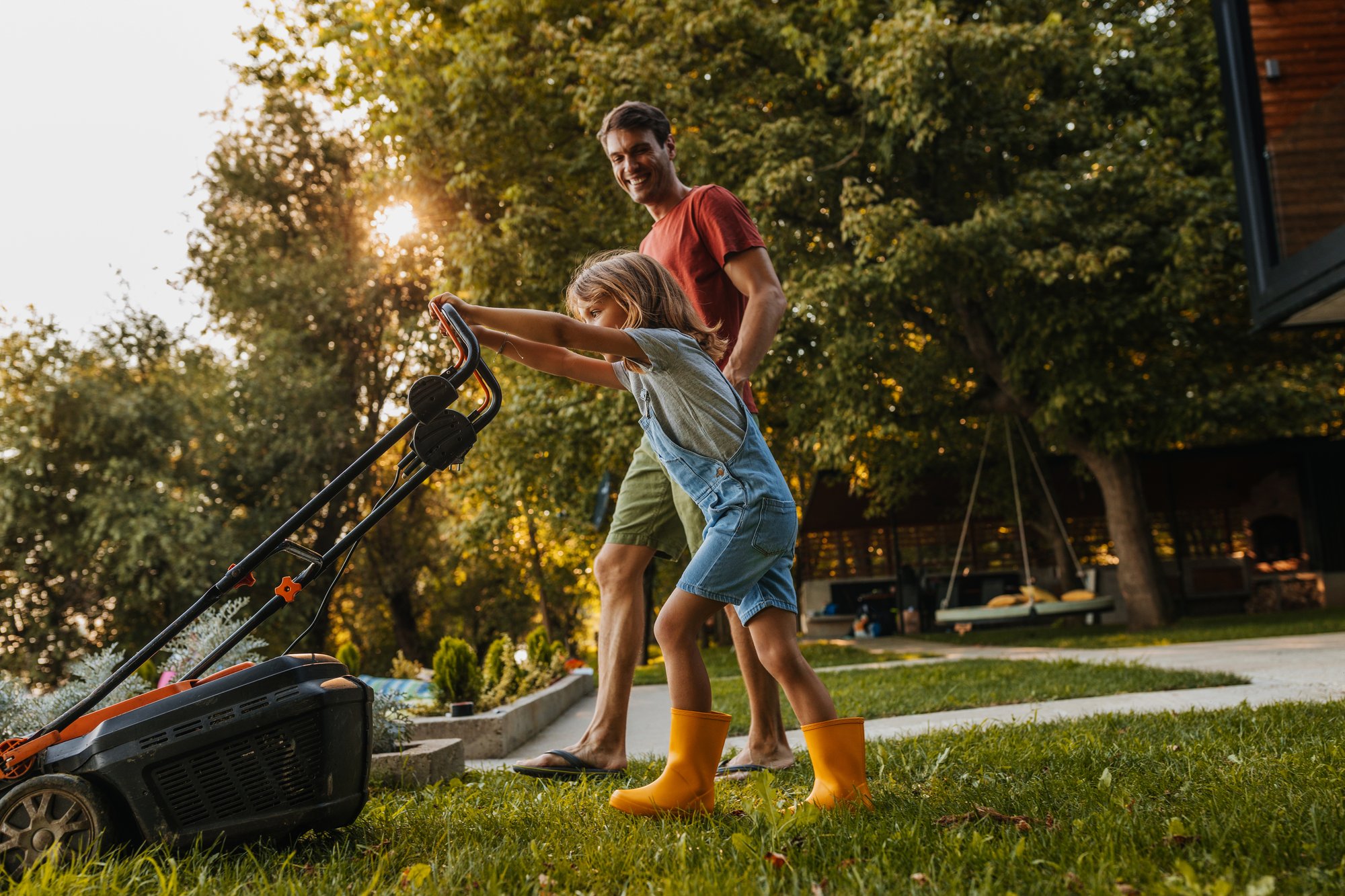 An adult watching his child with a smile who is mowing the lawn using a rented lawn mower.